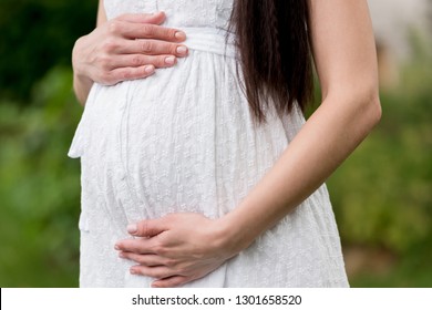 Mid Section Of Pregnant Woman In White Dress Touching Belly While Standing In Park 