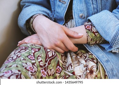 Mid Section Of Young Woman Wearing Denim Jacket Over Summer Dress With Floral Pattern - Close-up Of Female Hands Folded In Her Lap