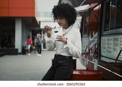 Mid Shot Of Mixed Race Young Woman With Afro Hair Holding Cardboard Box With Street Food And Eats It With Chopsticks. Food Truck At The Background