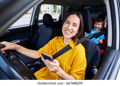 Mid Shot Of A Mother Driving A Car, Buckled The Seat Belt, Holding Phone. Laughing Boy On Back Seat