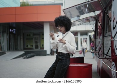 Mid Shot Of Pretty Ethnic Black Woman Having Asian Noodles From Cardboard Box For Lunch Outdoors Near Food Truck During Lunch Break. 