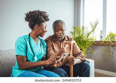 A mid-adult Black nurse in scrubs attentively assists an elderly Black man with his medication, highlighting compassionate in-home care in a cozy living room setting. - Powered by Shutterstock