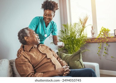 A mid-adult black nurse in scrubs smiles as she takes care of a senior black male, providing comfort and medical assistance in a warm living room setting. - Powered by Shutterstock