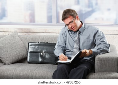 Mid-adult businessman taking notes into organiser while speaking on mobile phone sitting on sofa. - Shutterstock ID 49387255