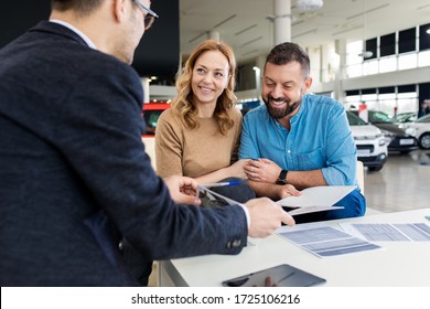 Mid-adult Couple Talking With Sales Agent And Signing Contract For New Car In Modern Car Showroom.