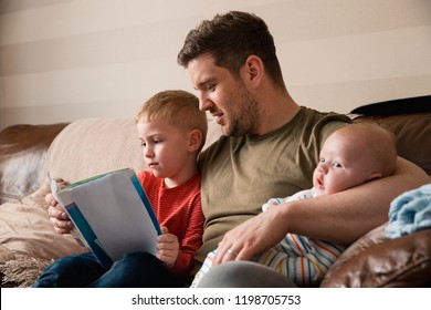 Mid-adult father is holding his newborn baby boy in his arm and then reading though the book with his son under the other arm. They all all sitting on the sofa in the living room. - Powered by Shutterstock