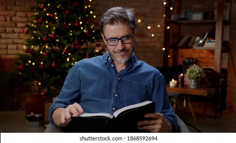 Mid-adult Older In Glasses Man Reading Book At Home At Christmas In Winter, Dark Room With Christmas Tree.