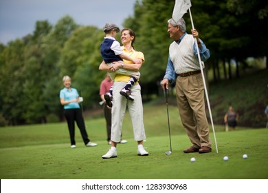Mid-adult woman carrying her young son while playing golf with her mature father and family.