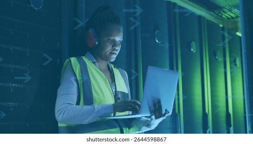 Mid-adult woman technician wearing vest examining racks in server room aisle, holding laptop. Technician, server, technology, professional, industrial, cybersecurity, modern