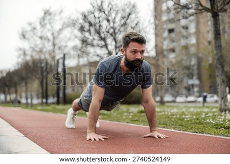 Mid-aged athletic man with dark beard performing push-ups on red running track. Wearing grey athletic shirt during outdoor training session, urban residential buildings visible in background.