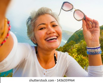 middle age 50s happy and cheerful Asian woman with grey hair taking selfie with mobile phone at beautiful tropical beach island smiling at cliff viewpoint enjoying Summer holidays travel destination - Powered by Shutterstock