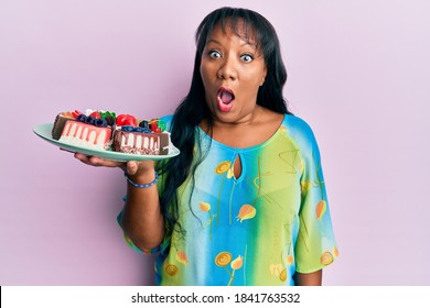 Middle age african american woman holding plate with cake slices scared and amazed with open mouth for surprise, disbelief face 