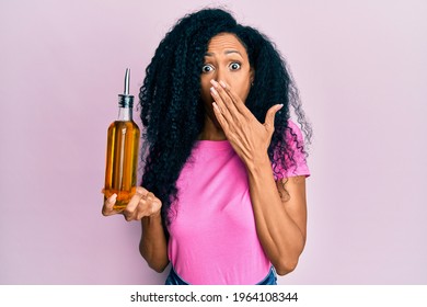 Middle Age African American Woman Holding Olive Oil Can Covering Mouth With Hand, Shocked And Afraid For Mistake. Surprised Expression 