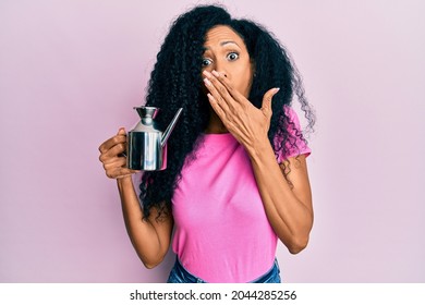 Middle Age African American Woman Holding Olive Oil Can Covering Mouth With Hand, Shocked And Afraid For Mistake. Surprised Expression 