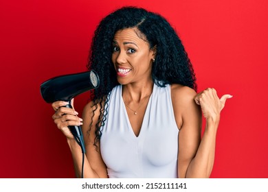 Middle Age African American Woman Holding Hair Dryer Pointing Thumb Up To The Side Smiling Happy With Open Mouth 