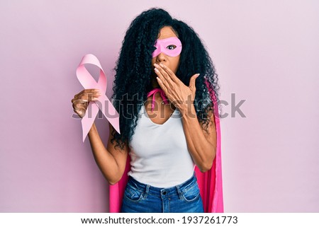 Middle age african american woman wearing super hero costume holding pink cancer ribbon covering mouth with hand, shocked and afraid for mistake. surprised expression 