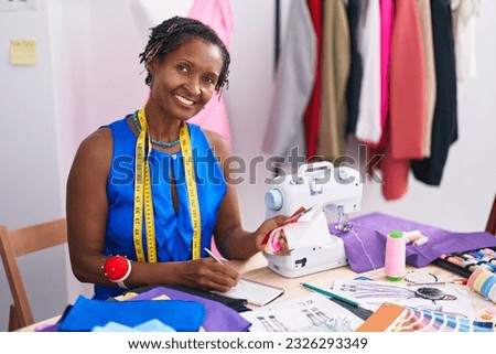 Middle age african american woman tailor writing on notebook holding cloth at atelier