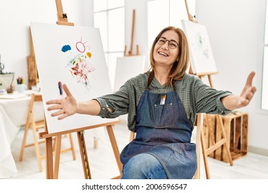 Middle age artist woman at art studio looking at the camera smiling with open arms for hug. cheerful expression embracing happiness. 