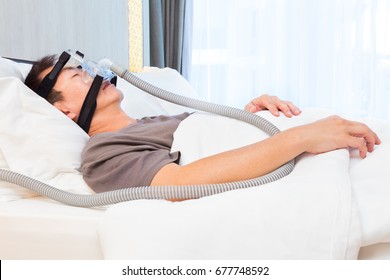 Middle Age Asian Man Sleeping In His Bed Wearing CPAP Mask Connecting To Air Hose, Device For People With Sleep Apnea