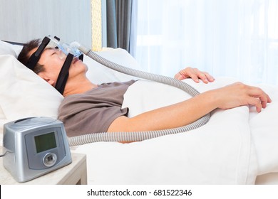 Middle Age Asian Man Sleeping In His Bed Wearing CPAP Mask Connecting To Air Hose And CPAP Machine, Device For People With Sleep Apnea