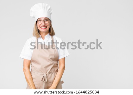 middle age baker woman looking happy and goofy with a broad, fun, loony smile and eyes wide open against flat wall