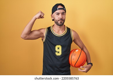 Middle Age Bald Man Holding Basketball Ball Over Yellow Background Strong Person Showing Arm Muscle, Confident And Proud Of Power 