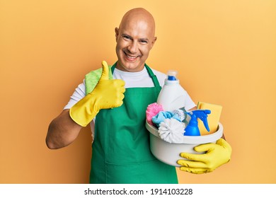 Middle age bald man wearing cleaner apron holding cleaning products smiling happy and positive, thumb up doing excellent and approval sign 