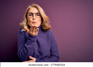 Middle age beautiful blonde woman wearing casual purple turtleneck sweater and glasses looking at the camera blowing a kiss with hand on air being lovely and sexy. Love expression.