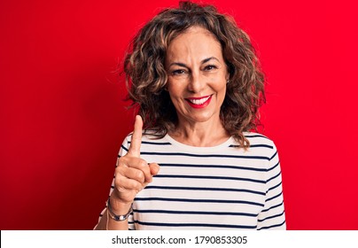 Middle age beautiful brunette woman wearing striped t-shirt standing over red background smiling with an idea or question pointing finger up with happy face, number one