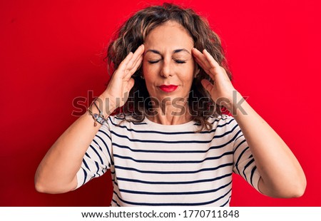 Middle age beautiful brunette woman wearing striped t-shirt standing over red background with hand on head, headache because stress. Suffering migraine.