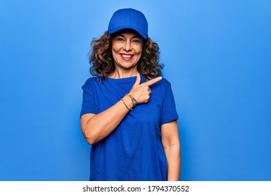 Middle age beautiful delivery woman wearing blue uniform and cap over isolated background smiling cheerful pointing with hand and finger up to the side
