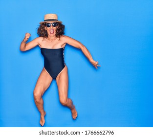 Middle Age Beautiful Hispanic Woman On Vacation Wearing Swimsuit,  Sunglasses And Hat Smiling Happy. Jumping With Smile On Face Celebrating Over Isolated Blue Background