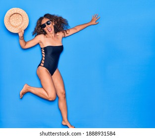 Middle Age Beautiful Hispanic Woman On Vacation Wearing Swimsuit,  Sunglasses And Hat Smiling Happy. Jumping With Smile On Face Over Isolated Blue Background