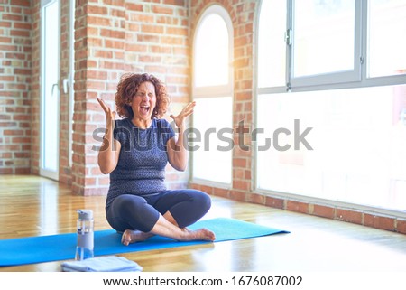 Middle age beautiful sportswoman wearing sportswear sitting on mat practicing yoga at home celebrating mad and crazy for success with arms raised and closed eyes screaming excited. Winner concept