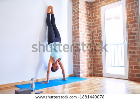 Middle age beautiful sportwoman smiling happy on mat. Practicing yoga doing wall-assisted handstand pose at gym