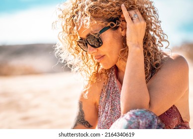 Middle age beautiful woman with curly hair and sunglasses in outdoor side portrait. Modern cute female people with tattoo relax at the beach under the summer sun. Healthy blonde people life - Powered by Shutterstock