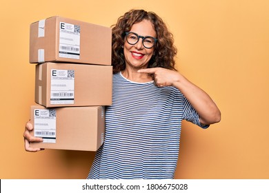 Middle age beautiful woman holding packages with label over isolated yellow background smiling happy pointing with hand and finger