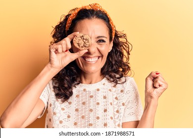 Middle age beautiful woman holding chocolate cookie over eye screaming proud, celebrating victory and success very excited with raised arm 