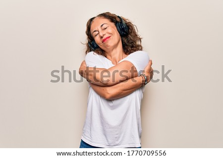 Middle age beautiful woman listening to music using headphones over white background hugging oneself happy and positive, smiling confident. Self love and self care
