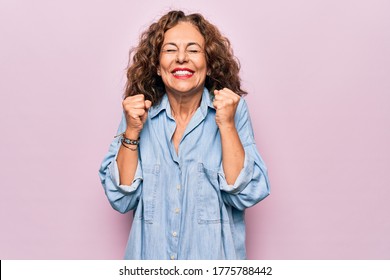 Middle age beautiful woman wearing casual denim shirt standing over pink background excited for success with arms raised and eyes closed celebrating victory smiling. Winner concept. - Powered by Shutterstock