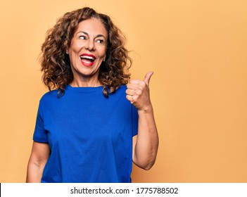 Middle age beautiful woman wearing casual t-shirt standing over isolated yellow background pointing thumb up to the side smiling happy with open mouth