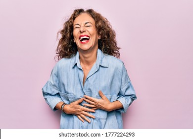 Middle age beautiful woman wearing casual denim shirt standing over pink background smiling and laughing hard out loud because funny crazy joke with hands on body. - Powered by Shutterstock