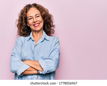 Eine schöne Frau im Mittelalter, die ein lässiges Denim-Hemd trägt, das auf rosafarbenem Hintergrund steht, und ein glückliches Gesicht, das mit gekreuzten Armen, die die Kamera anschauen, lächelt. Positive Person.