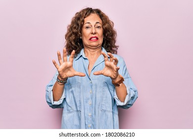 Middle age beautiful woman wearing casual denim shirt standing over pink background afraid and terrified with fear expression stop gesture with hands, shouting in shock. Panic concept.