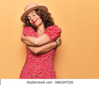 Middle age beautiful woman wearing casual dress and hat over isolated yellow background hugging oneself happy and positive, smiling confident. Self love and self care