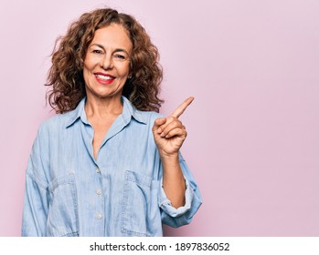 Middle age beautiful woman wearing casual denim shirt standing over pink background with a big smile on face, pointing with hand finger to the side looking at the camera.