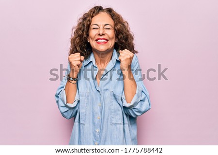 Middle age beautiful woman wearing casual denim shirt standing over pink background excited for success with arms raised and eyes closed celebrating victory smiling. Winner concept.
