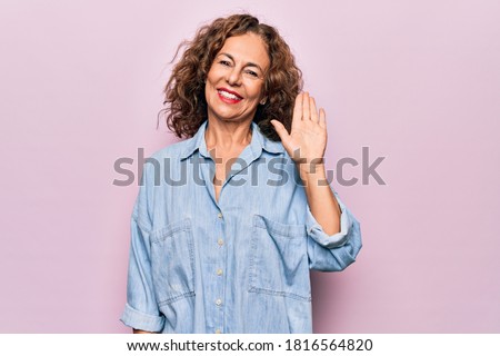 Middle age beautiful woman wearing casual denim shirt standing over pink background Waiving saying hello happy and smiling, friendly welcome gesture
