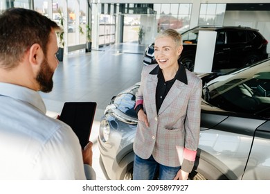 Middle Age Blond Cahrming Woman Smiling While Discussing Payment Rates For A New Car With A Car Salesman In An Automotive Dealership Salon.