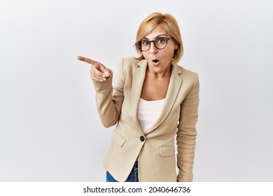 Middle age blonde business woman standing over isolated background surprised pointing with finger to the side, open mouth amazed expression. 